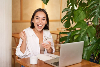 快乐的年轻的亚洲女人赢得庆祝胜利成就收到好新闻移动电话坐着移动PC工作