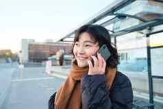 铣削朝鲜文女孩会说话的移动电话站公共汽车停止智能手机摆姿势路冬天包装围巾穿黑色的夹克