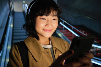 关闭可爱的亚洲女孩耳机选择首歌听通勤自动扶梯<strong>地铁</strong>微笑阅读消息移动<strong>电话</strong>