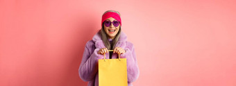 时尚亚洲高级女人购物太阳镜时尚的冬天衣服持有纸袋商店微笑快乐相机粉红色的背景