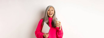 快乐亚洲高级女企业家笑喝咖啡持有移动PC站粉红色的毛衣白色背景