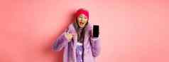 在线购物时尚概念很酷的亚洲奶奶时尚的人造皮毛外套指出手指左空白智能手机屏幕检查互联网促销粉红色的背景