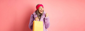 购物时尚概念快乐亚洲高级女人赢得持有纸袋使拳头泵手势快乐的脸站粉红色的背景
