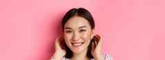 美皮肤护理概念特写镜头可爱的微笑亚洲女人挤进头发耳朵脸红盯着相机站粉红色的背景