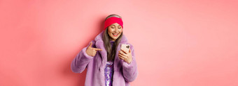 在线购物时尚概念很酷的亚洲高级女人检查促销活动移动电话指出手指智能手机屏幕微笑很高兴粉红色的背景