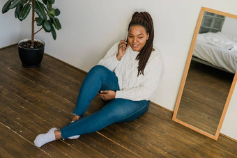 积极的年轻的非洲美国女人聊天手机微笑地板上首页快乐的年轻的女人沟通智能手机在室内