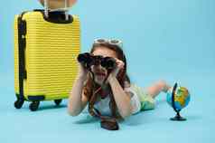 可爱的女孩双筒望远镜说谎全球黄色的手提箱蓝色的背景旅行概念