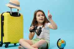 可爱的旅行者女孩点手指复制空间蓝色的背景坐着黄色的手提箱全球