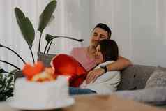 美丽的年轻的夫妇坐着沙发上拥抱浪漫的日期情人节一天首页妻子丈夫庆祝心形状气球蛋糕花