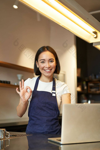 肖像咖啡师女孩亚洲女咖啡馆工人显示标志批准推荐smth快乐很高兴穿围裙站移动PC计数器