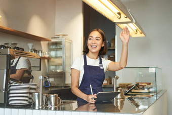 快乐亚洲女孩咖啡师提高手臂邀请咖啡馆客户订单咖啡站计数器回来终端平板电脑