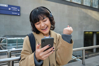 快乐的朝鲜文女孩赢了庆祝胜利智能手机应用程序胜利表达快乐兴奋站耳机空街