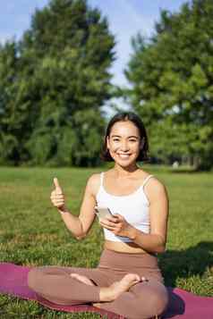 垂直拍摄亚洲女人显示拇指推荐瑜伽培训在线冥想应用程序练习新鲜的空气公园