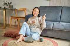 微笑亚洲女孩坐在地板上时尚的生活房间指出手指广告显示促销横幅持有移动电话手