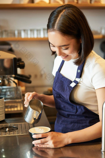 微笑亚洲女孩咖啡师咖啡馆工作人员倒蒸牛奶咖啡准备卡布奇诺咖啡拿铁艺术站蓝色的围裙计数器