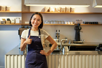 微笑女孩学生工作兼职咖啡馆咖啡师显示拇指穿围裙站咖啡商店计数器