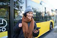 肖像女孩站公共汽车停止等待公共运输检查时间表智能手机应用程序持有移动电话穿温暖的衣服