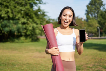 微笑亚洲健身女孩橡胶瑜伽席显示智能<strong>手机屏</strong>幕建议锻炼应用程序站草坪上公园