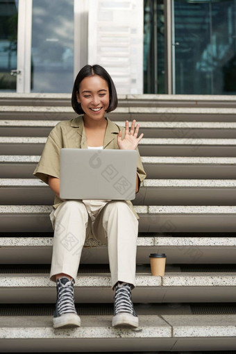 肖像年轻的亚洲女孩<strong>学生会</strong>谈移动PC视频闲谈，聊天说话在线会议坐着在户外楼梯