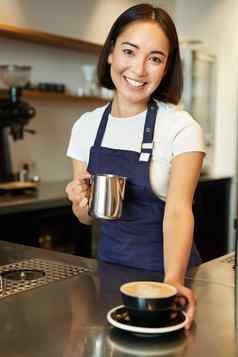 垂直拍摄微笑亚洲女孩咖啡馆给卡布奇诺咖啡使订单准备咖啡站计数器蓝色的围裙