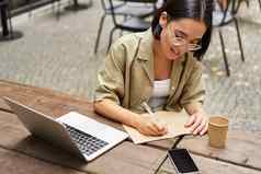 肖像年轻的女人研究在线坐着移动PC写作使笔记电脑屏幕坐着咖啡馆在户外
