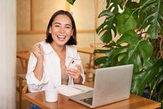快乐的年轻的亚洲女人赢得庆祝胜利成就收到好新闻移动电话坐着移动PC工作