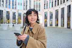 时尚的朝鲜文女孩耳机听音乐移动电话站城市中心等待街写文本消息智能手机