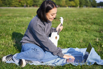 微笑亚洲女孩<strong>学习</strong>玩尤克里里琴移动PC在线<strong>视频教程</strong>坐着草公园音乐的仪器