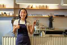 快乐年轻的亚洲咖啡师女工作人员工人围裙显示拇指指出复制空间展示咖啡馆广告