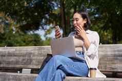 亚洲女孩惊讶智能手机屏幕检查移动电话通知兴奋坐着移动PC公园板凳上