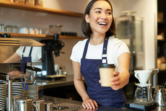 微笑亚洲咖啡师女孩给外卖咖啡杯准备外卖订单客人咖啡馆穿围裙统一的