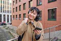 年轻的快乐女人沟外套听音乐耳机持有智能手机移动应用程序找到路线城市站街背包