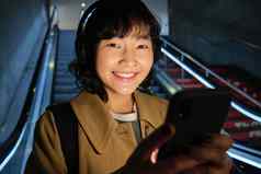 关闭可爱的亚洲女孩耳机选择首歌听通勤自动扶梯地铁微笑阅读消息移动电话