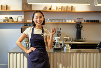 肖像亚洲女孩围裙站咖啡馆计数器咖啡师指出手指复制空间空横幅咖啡商店图片