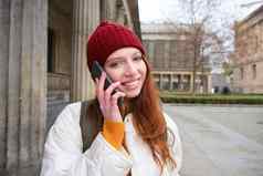 微笑年轻的红色头发的人女人听的声音消息使电话调用走街会谈智能手机