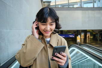 肖像可爱的朝鲜文女孩沟外套自动扶梯持有移动<strong>电话</strong>微笑很高兴<strong>手表</strong>视频无线耳机