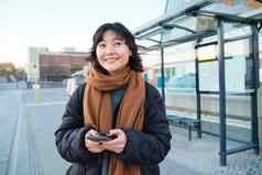 快乐微笑朝鲜文女孩移动电话站公共汽车停止智能手机离开时间表应用程序摆姿势冬天衣服