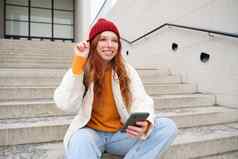 时尚的欧洲女孩红色的头发坐在公共楼梯智能手机的地方在线订单发送消息移动电话社会应用程序微笑幸福的
