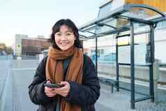快乐微笑朝鲜文女孩移动电话站公共汽车停止智能手机离开时间表应用程序摆姿势冬天衣服