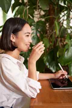 美丽的亚洲女孩坐着咖啡馆杯咖啡画数字平板电脑图形笔轻而易举的事使scatches