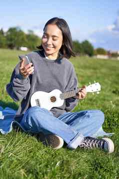 垂直拍摄微笑亚洲女孩智能手机玩尤克里里琴阅读和弦歌词唱歌放松在户外生活方式人概念