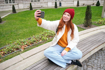 年轻的青少年红色头发的人女孩坐在板凳上公园需要自拍使照片智能手机应用程序记录视频博客