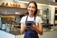 微笑亚洲女孩咖啡师使咖啡持有杯穿咖啡馆统一的围裙站计数器