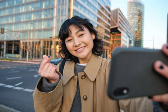 积极的亚洲女孩需要自拍智能手机显示心手指使照片街旅游摆姿势移动<strong>电话</strong>