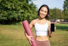 微笑亚洲健身女孩橡胶瑜伽席显示智能手机屏幕建议锻炼应用程序站草坪上公园