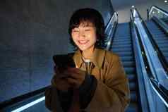 微笑朝鲜文女孩自动扶梯黑暗持有移动电话智能手机应用程序听音乐通勤城市