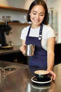 垂直拍摄微笑亚洲女孩咖啡馆给卡布奇诺咖啡使订单准备咖啡站计数器蓝色的围裙
