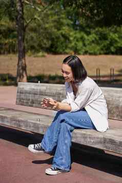 快乐微笑亚洲女孩坐着板凳上公园浏览社会媒体持有智能手机移动电话应用程序