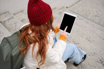 后视图红色头发的人女孩触摸数字平板电脑屏幕触控板文本消息互联网应用程序小工具坐在楼梯在户外