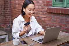微笑亚洲女人移动PC无线耳机支付信贷卡购买在线坐着杯咖啡